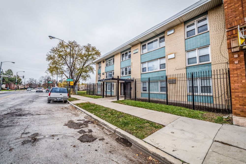 Apartment building with blue accents, metal fence, and sidewalk. Cloudy day.