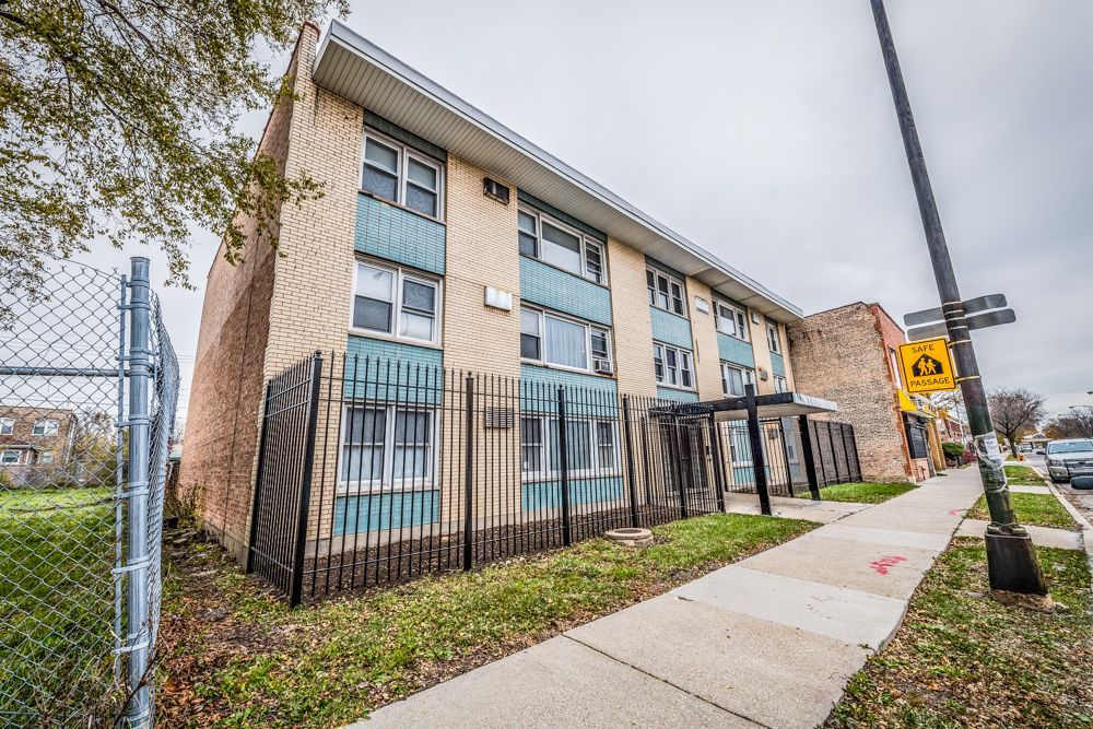 Three-story apartment building with metal fence, sidewalk, and street sign on a cloudy day.