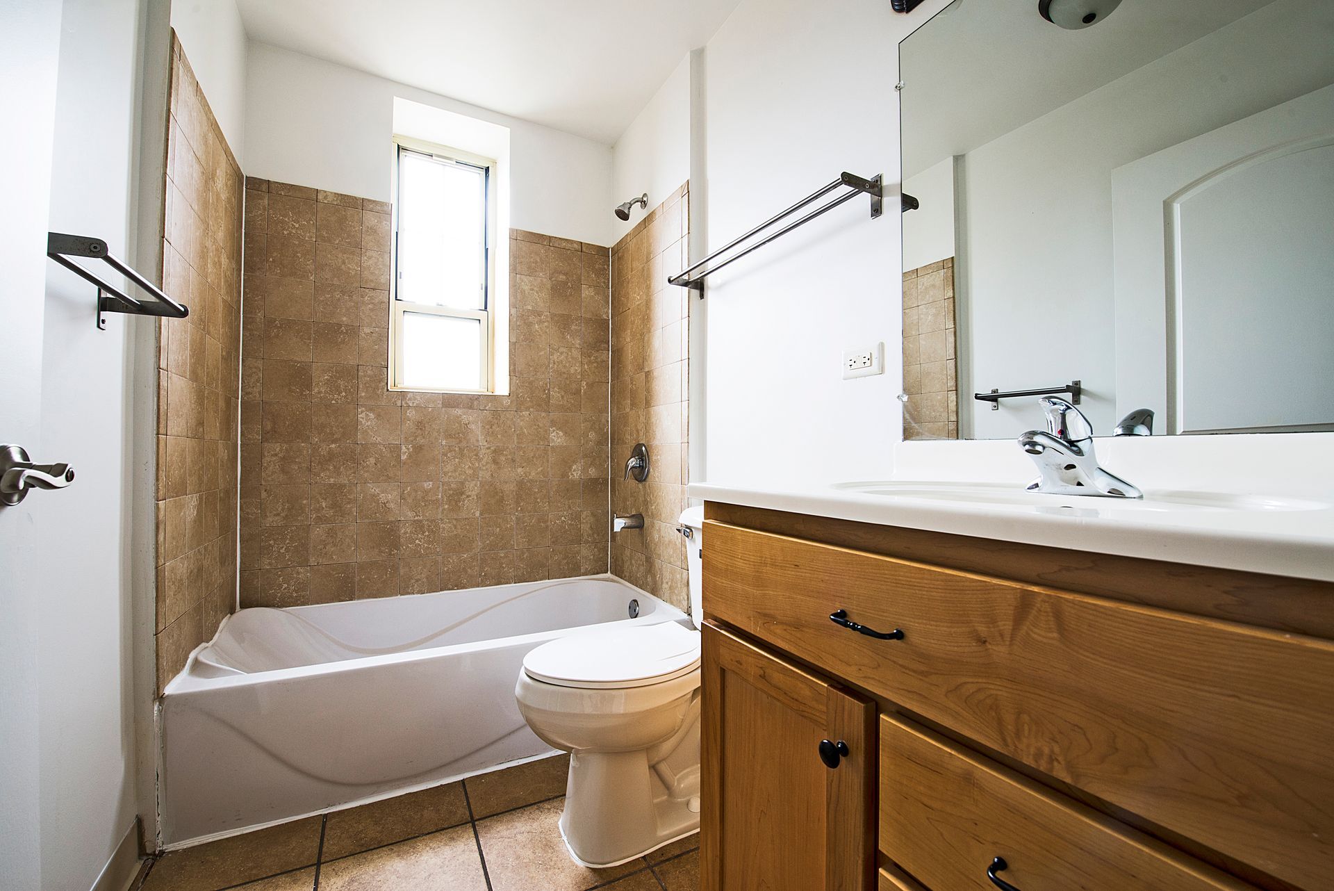 Bathroom with beige tiled walls, white bathtub and toilet, light wood vanity, and a window.