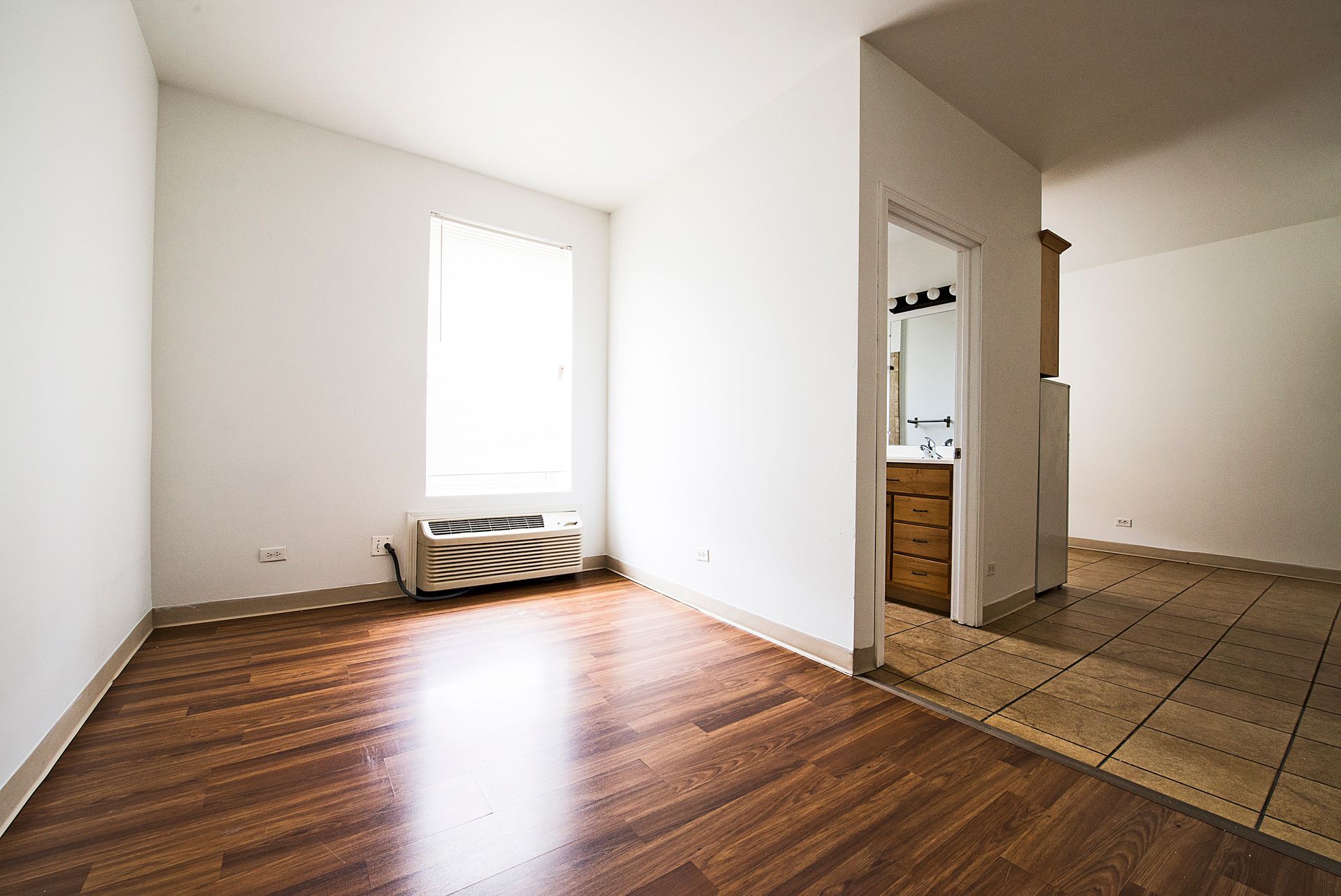 Empty room with wood floors and a bathroom visible through a doorway. A window with blinds and an air conditioner are present.