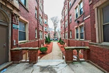 A red brick apartment complex with a walkway between buildings, trees, and cloudy sky.