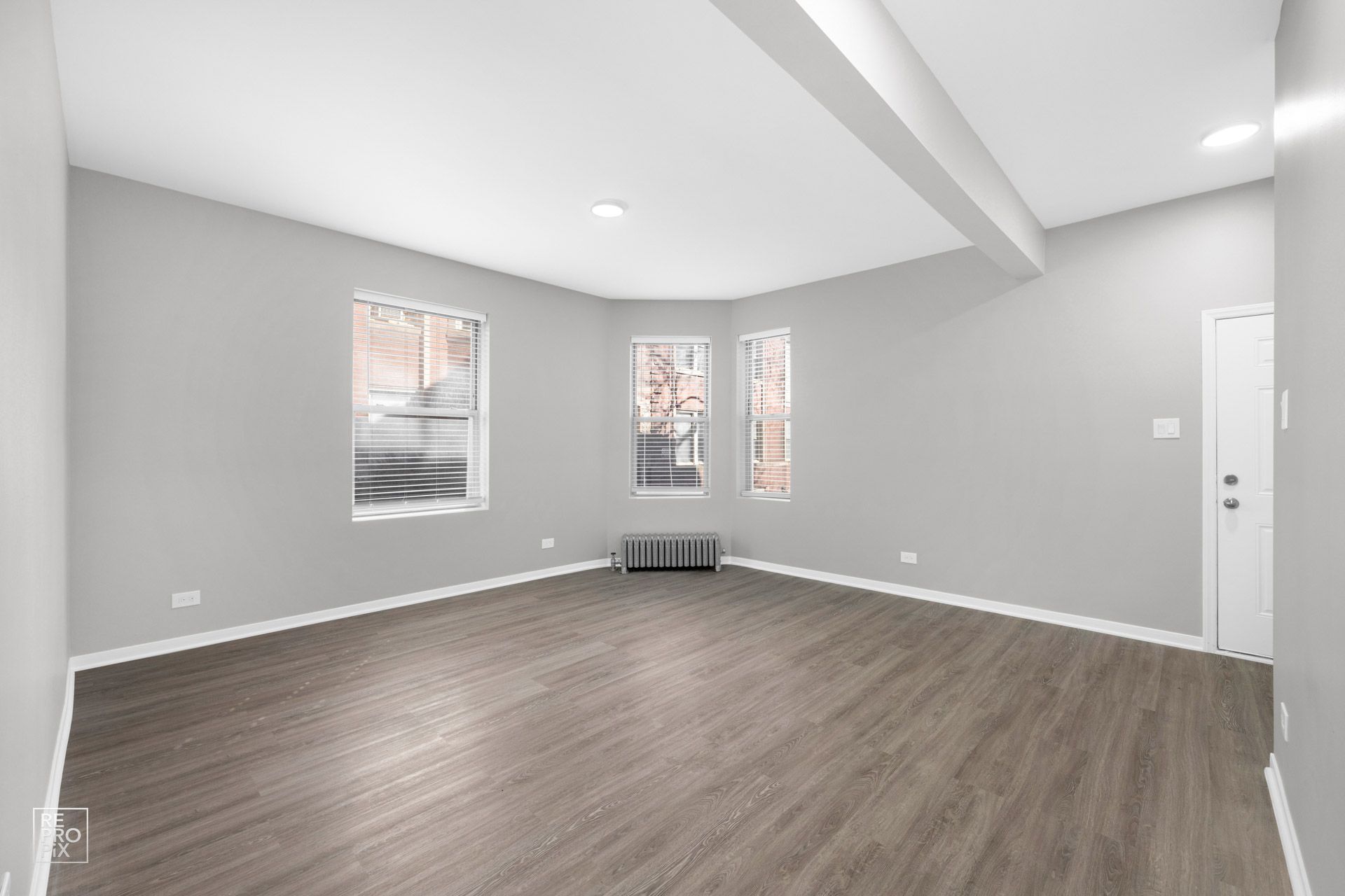Empty, gray-walled living room with three windows and dark wood-look flooring. White trim, recessed lighting.