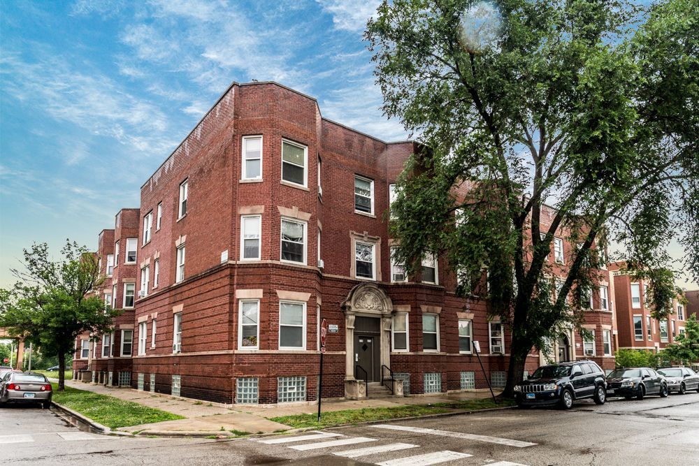 Red brick apartment building on a corner lot with a tree and cars parked on the street.