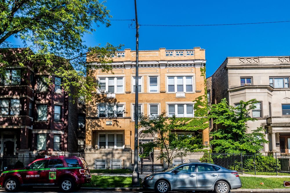 A three-story yellow brick building between two other buildings; cars parked in front, trees on either side.