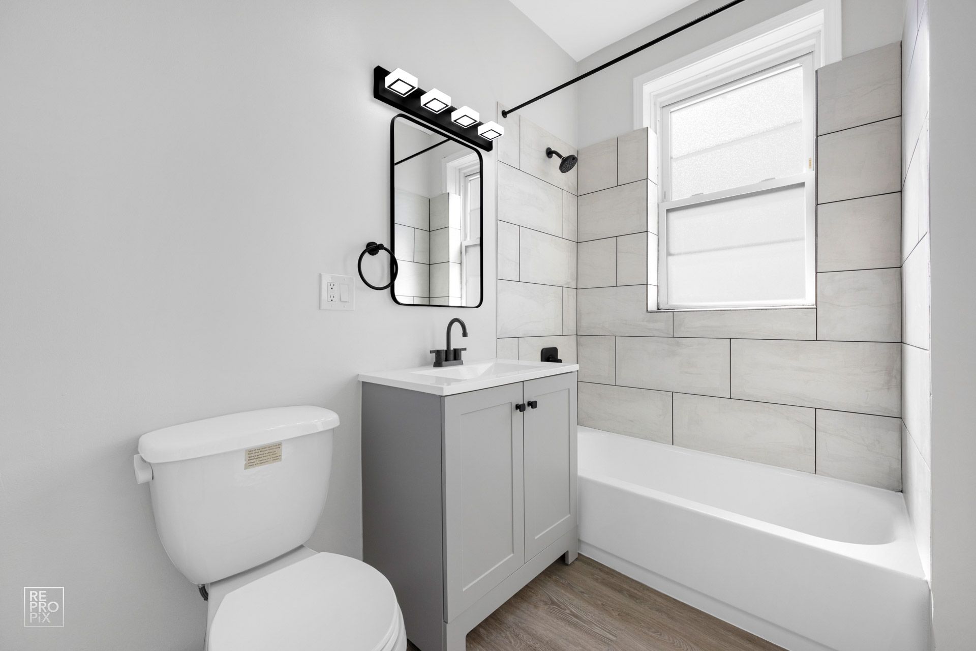 Bathroom with gray vanity, white toilet, tiled shower, and a window.