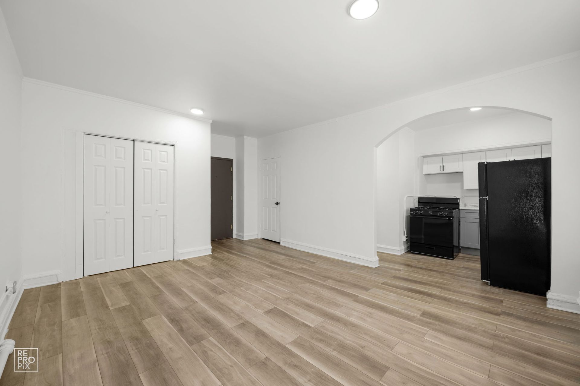 Empty, bright living space with wooden floors, white walls, and a view into the kitchen.