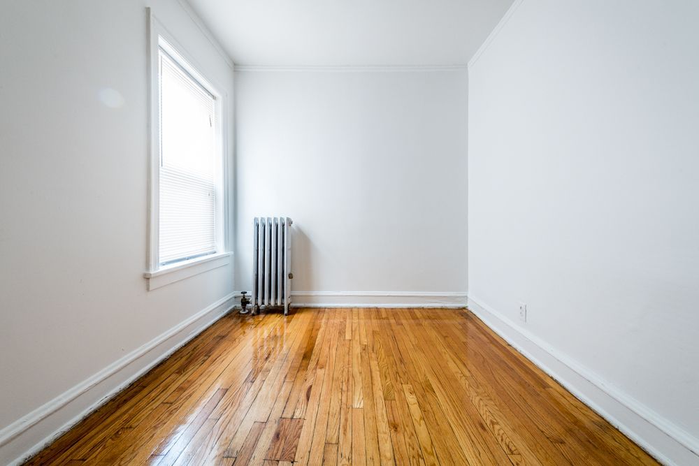 Empty room with wood floor, radiator, and window. White walls, bright natural light.