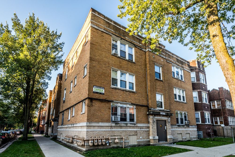 Brick apartment building with multiple windows; sidewalk and trees in front.