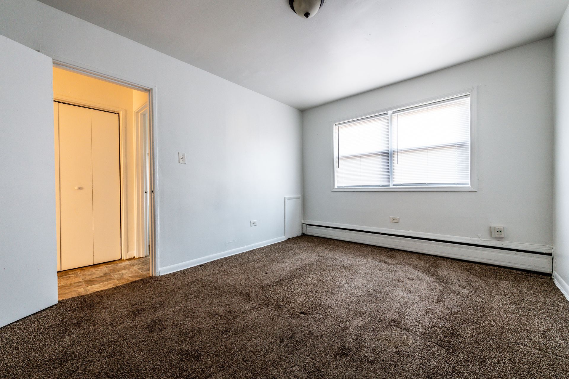 Empty room with white walls, brown carpet, window with blinds, and closet door.