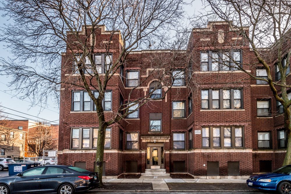Brick apartment building with multiple windows, trees in front, and cars parked on the street.