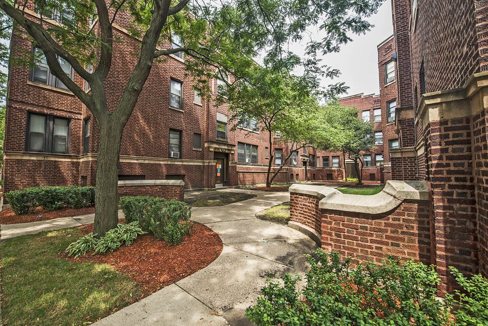 Brick apartment buildings with a courtyard, trees, bushes, and concrete pathways.