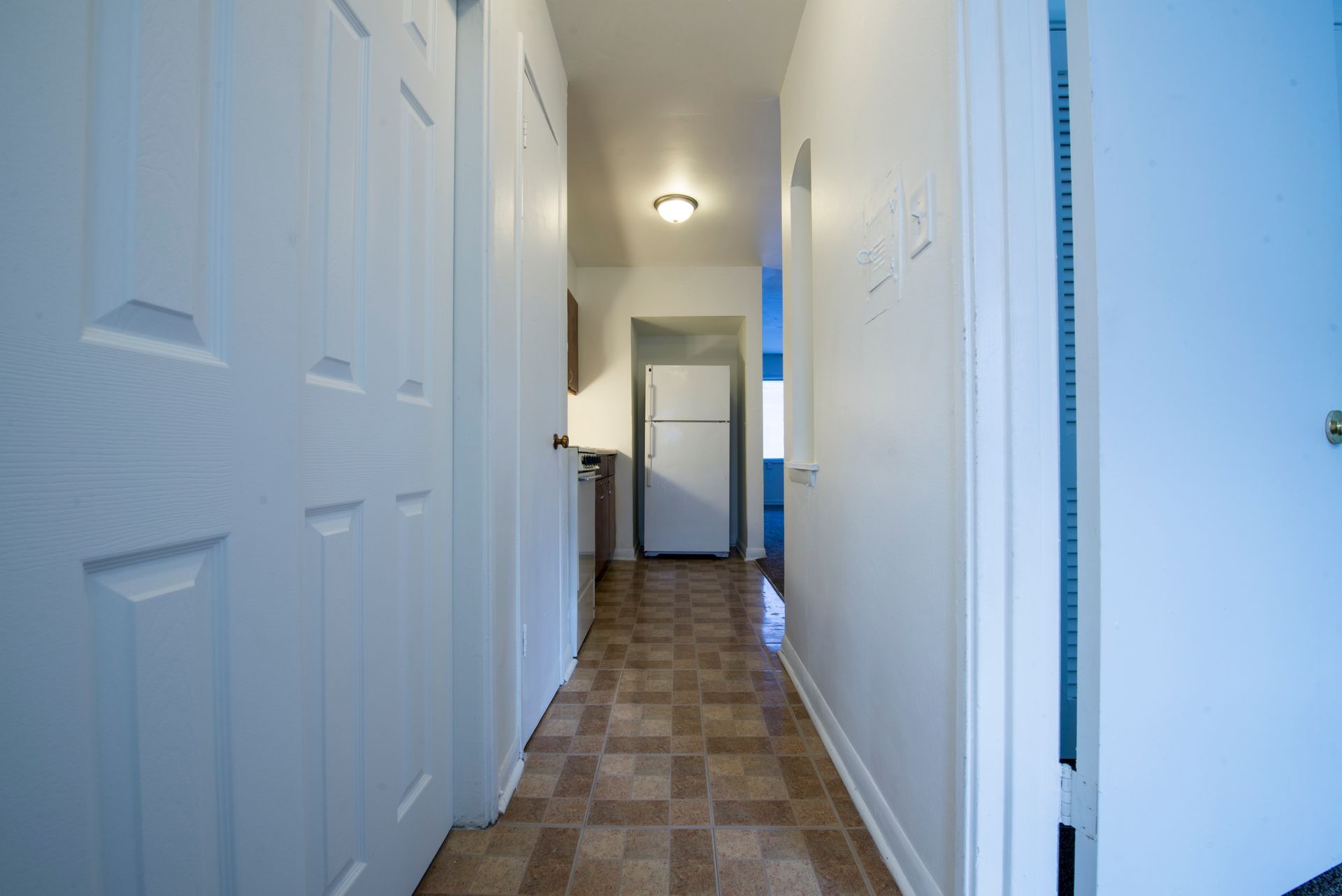 Narrow hallway with white doors and speckled floor leading to a kitchen with a refrigerator.