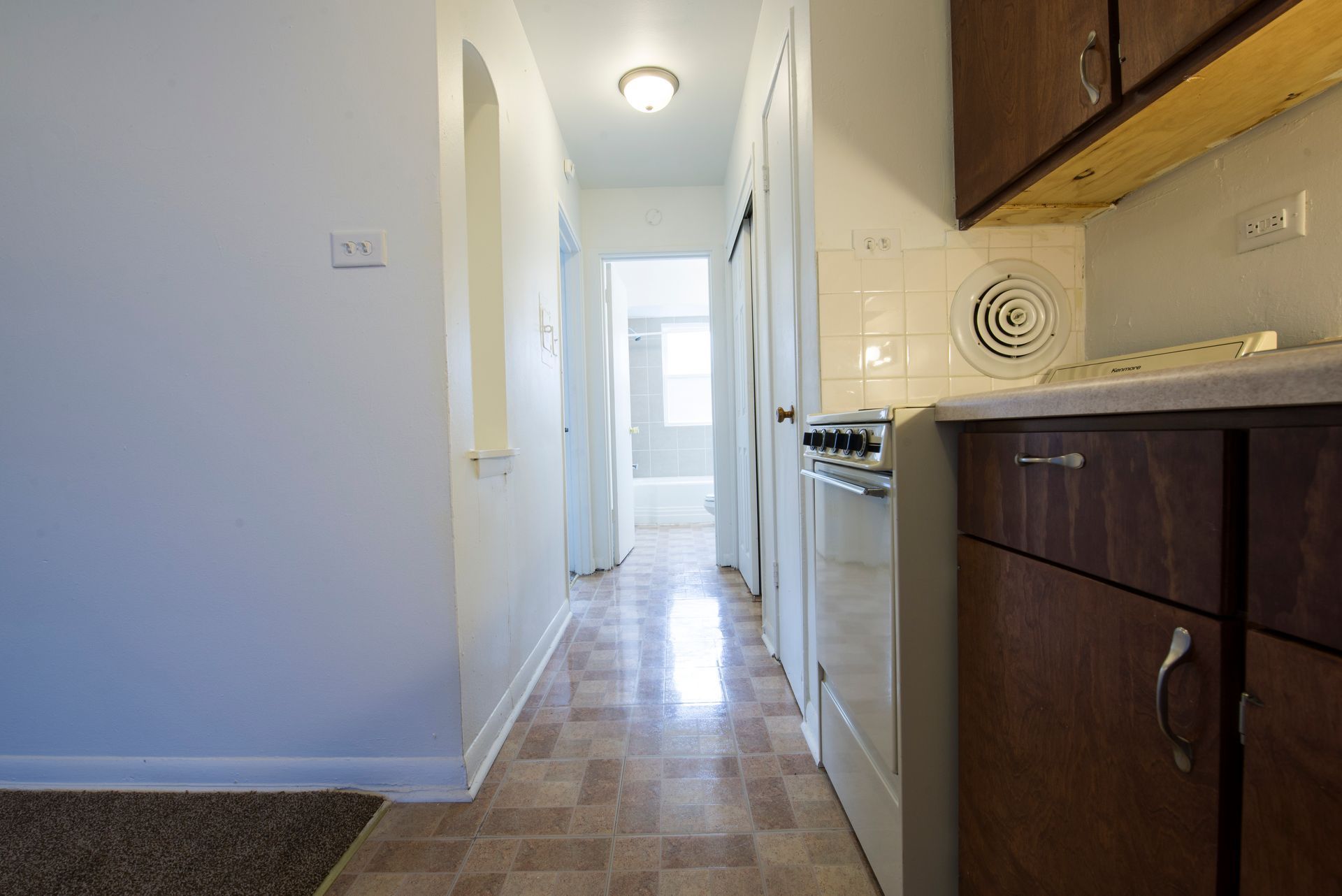 Narrow apartment hallway leads to bathroom. Kitchen on right with cabinets, stove, and fan.