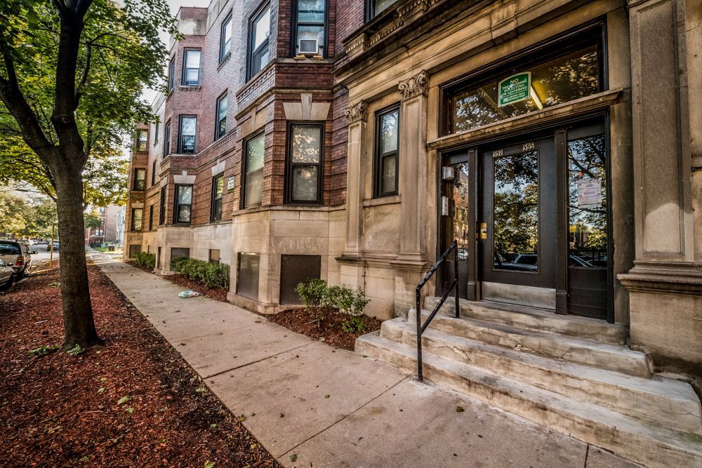 Apartment building with stone facade, dark doors, steps, and sidewalk. Brown brick, green tree, cars parked on street.