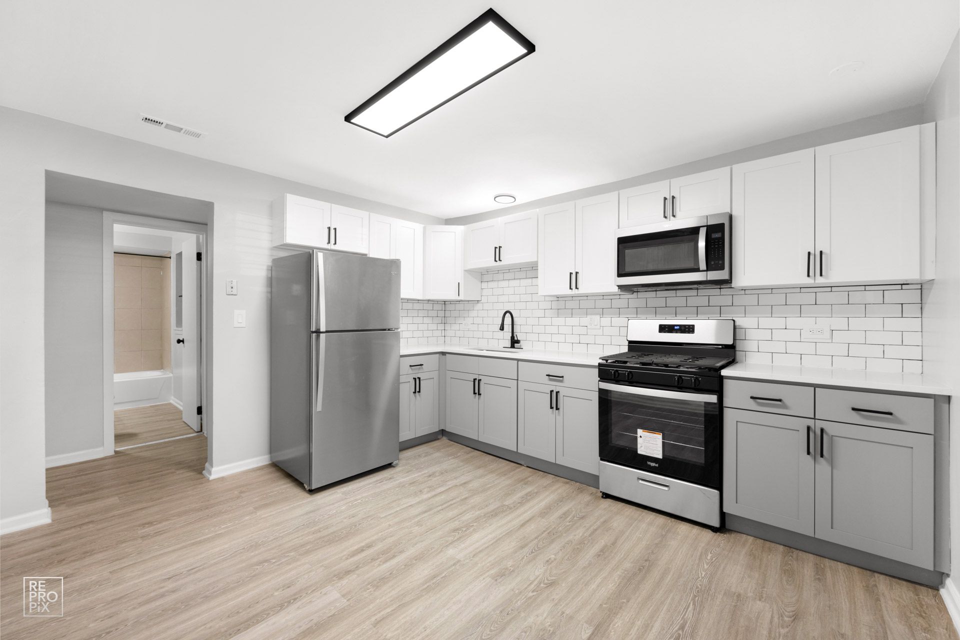 Gray and white kitchen with stainless steel appliances and wood-look flooring. Entryway to bathroom on the left.