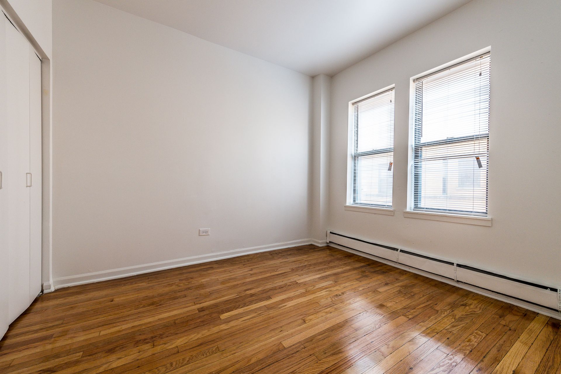 Empty room with hardwood floors, white walls, two windows with blinds, and a closet.