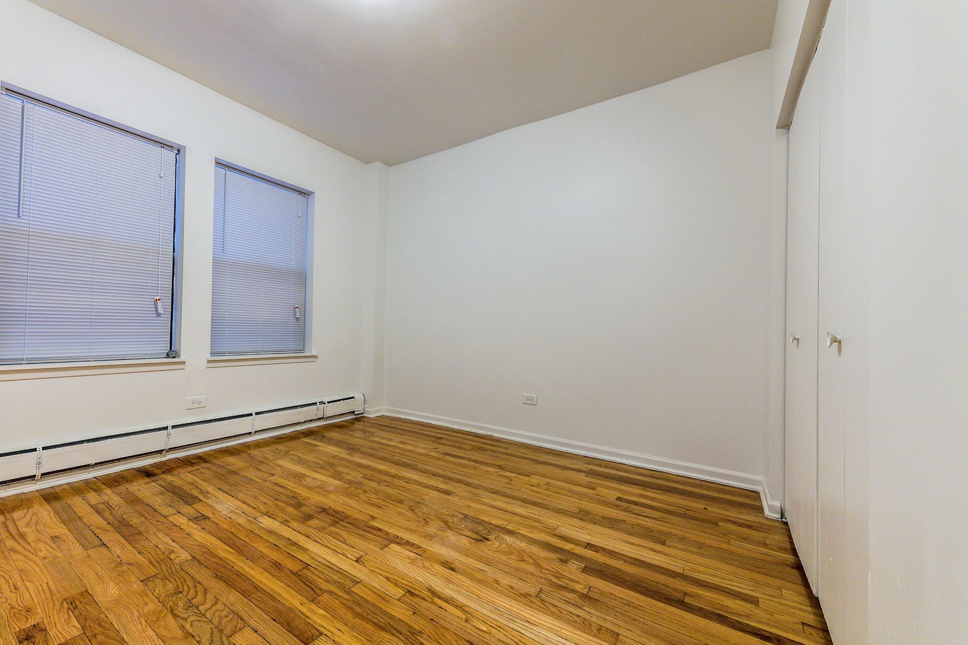 Empty room with hardwood floors, two windows with blinds, and a white closet.