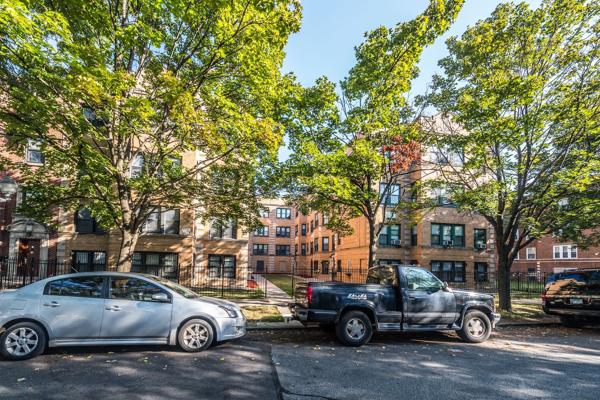 Cars parked on a street in front of brick apartment buildings shaded by green trees on a sunny day.