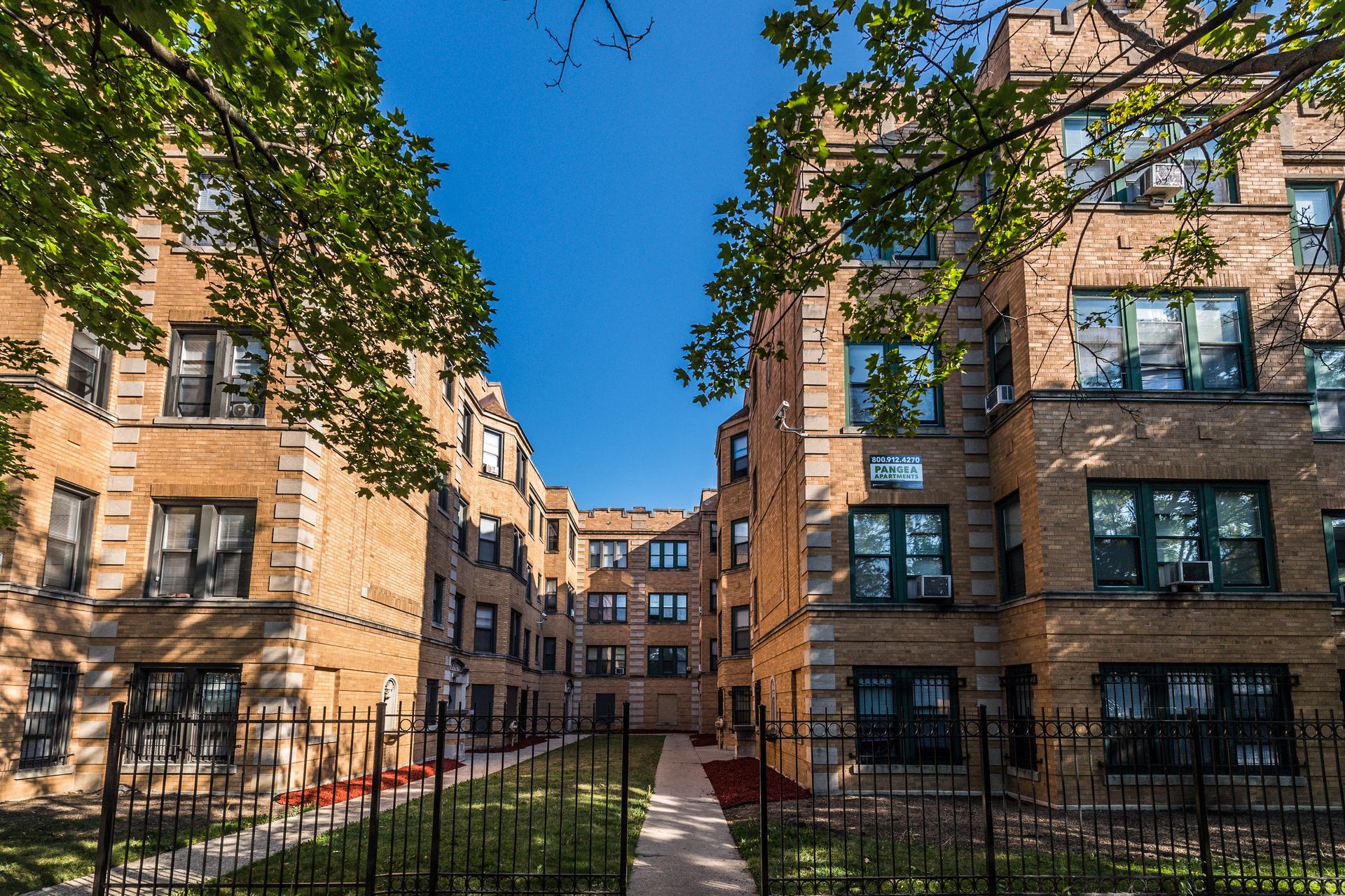 Apartment buildings with brick facades, black fence, green grass, and blue sky.