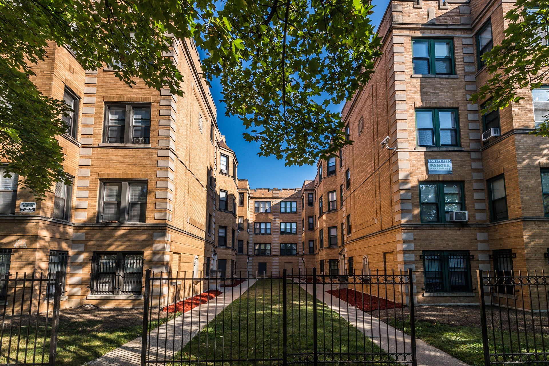 Brick apartment buildings with a courtyard and a wrought iron fence under a blue sky.