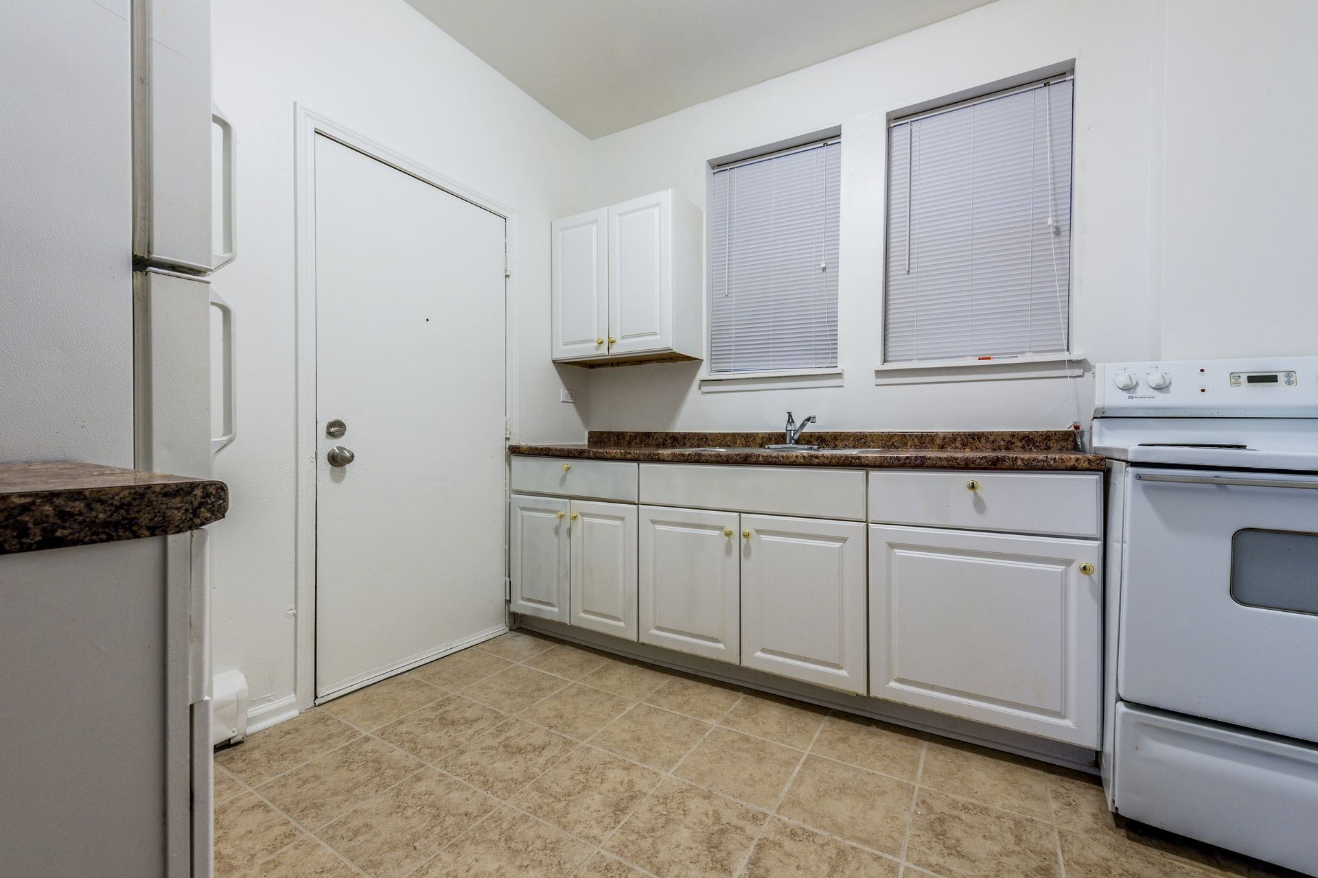 White kitchen with cabinets, stove, sink, refrigerator, and door. Brown countertops and beige tile floor.