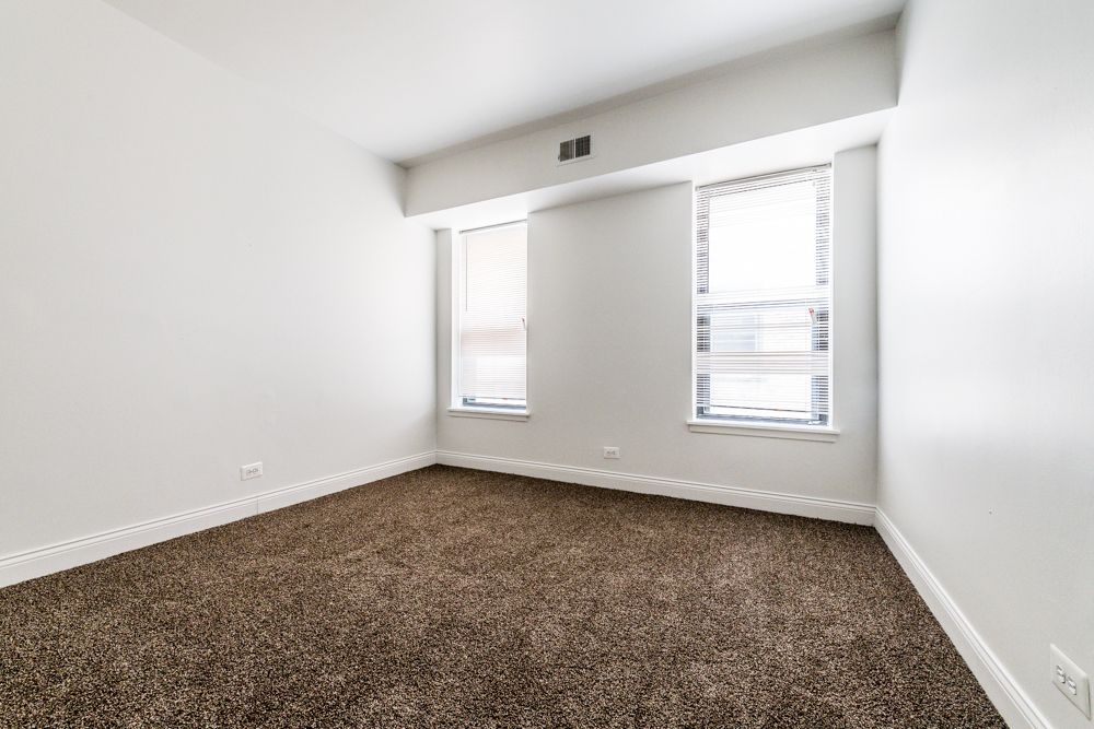Empty room with brown carpet, two windows, and white walls.
