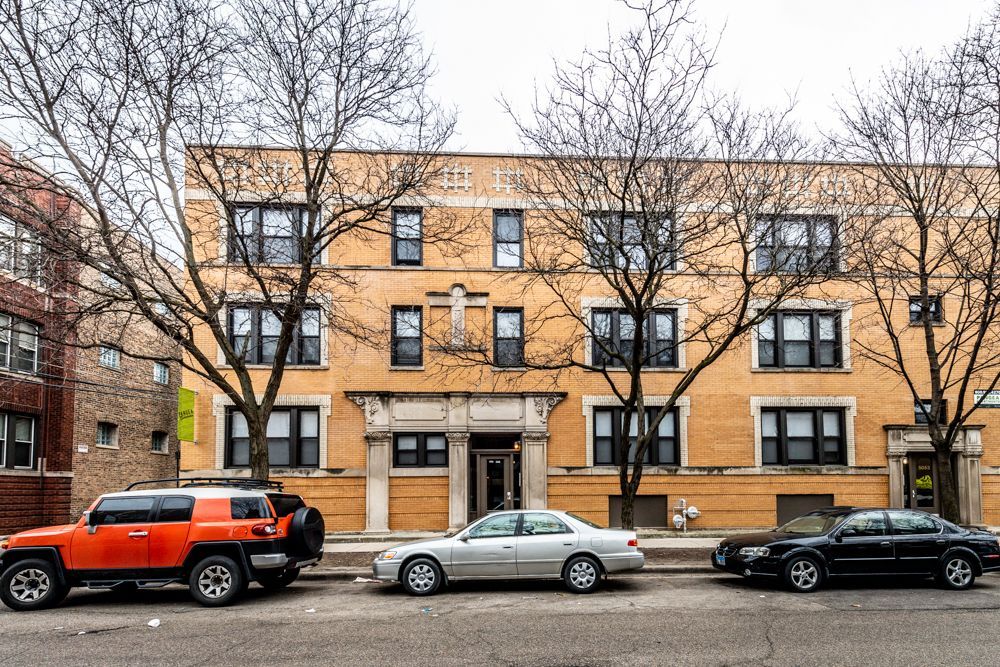 Three-story yellow brick apartment building with parked cars in front and bare trees.