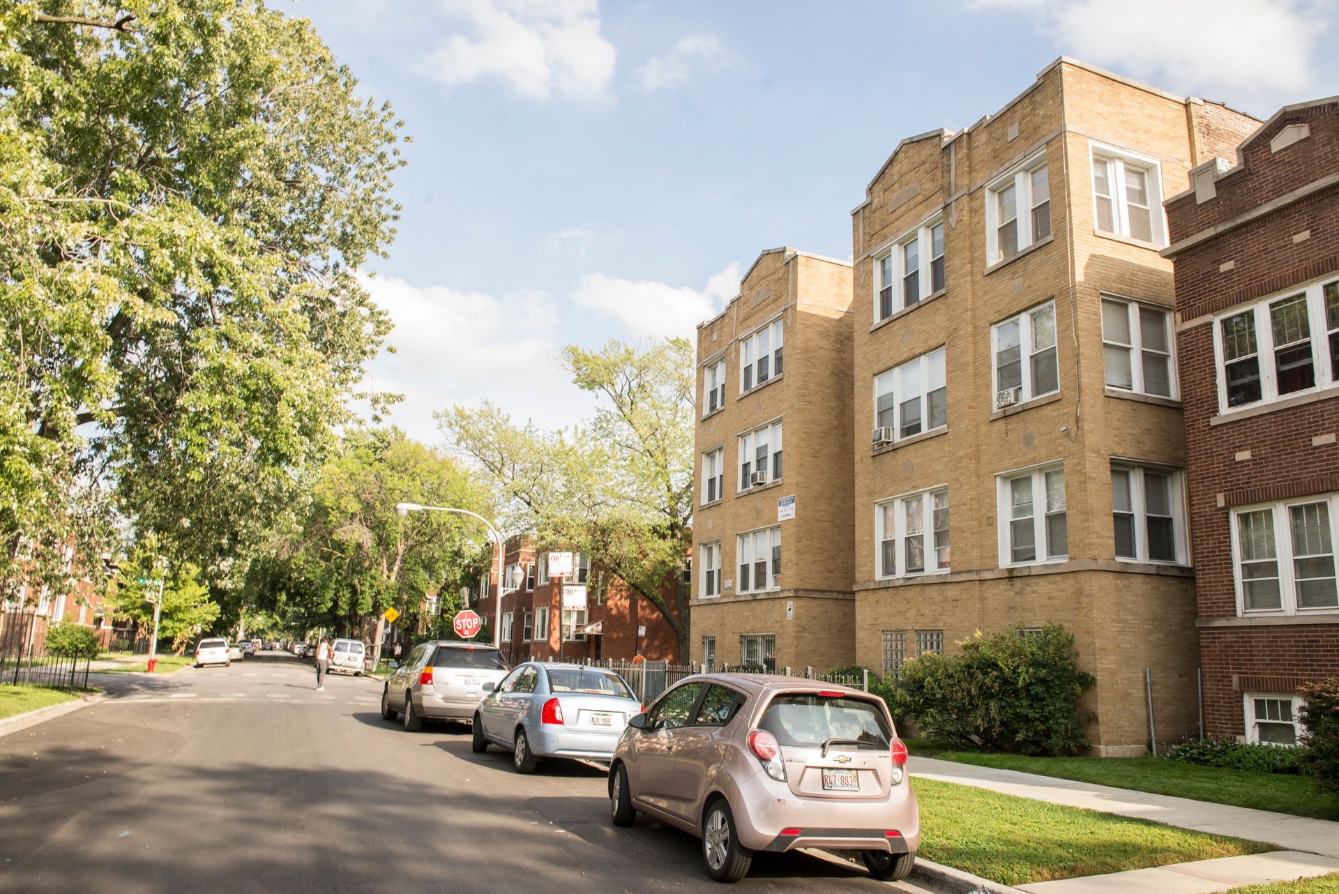 Street with parked cars in front of three-story brick apartment buildings, sunny day.