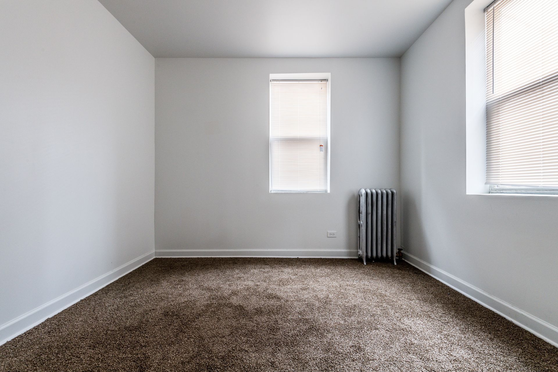Empty room with beige carpet, white walls, two windows with blinds, and a radiator.