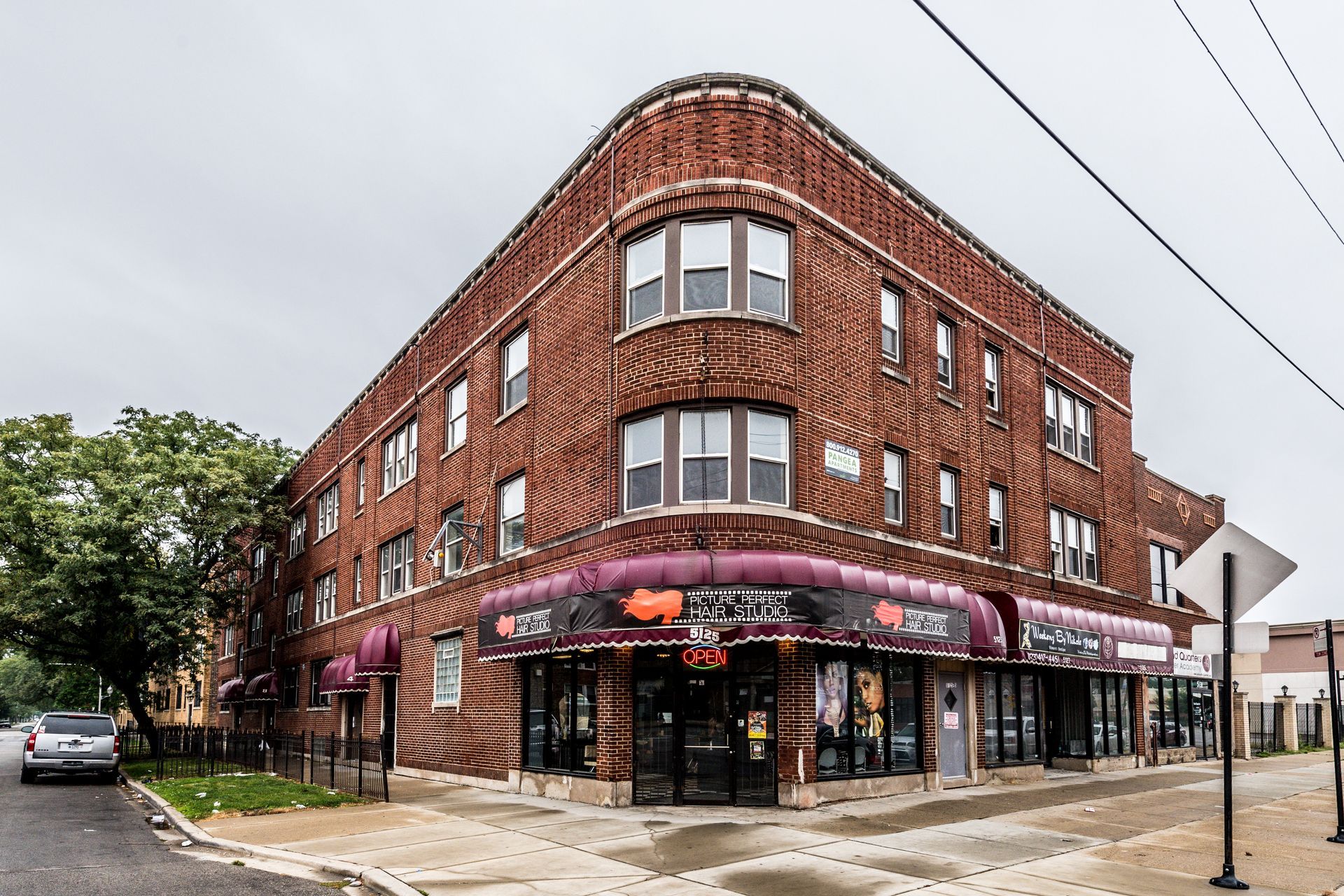 Brick building on a corner lot, with storefronts below apartments. Purple awnings, cloudy sky.