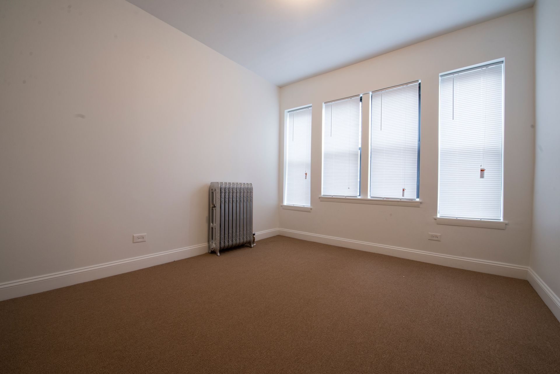 Empty room with brown carpet, three windows with blinds, and a radiator.