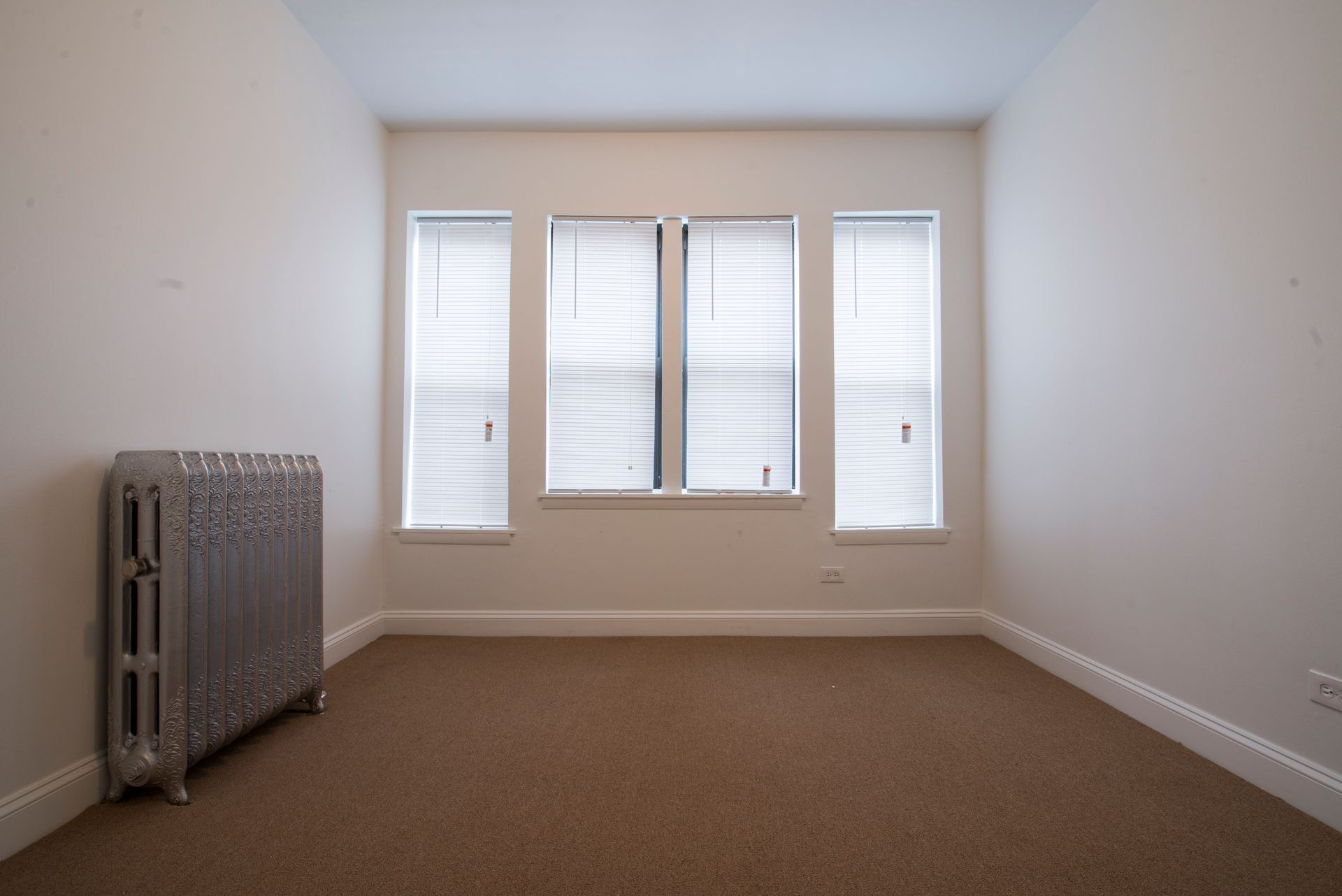 Empty room with a brown carpet, three windows, a radiator, and white walls.