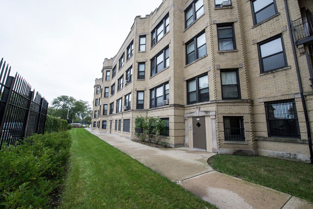 Apartment building exterior, tan brick, curved facade, sidewalk, grass, overcast sky.