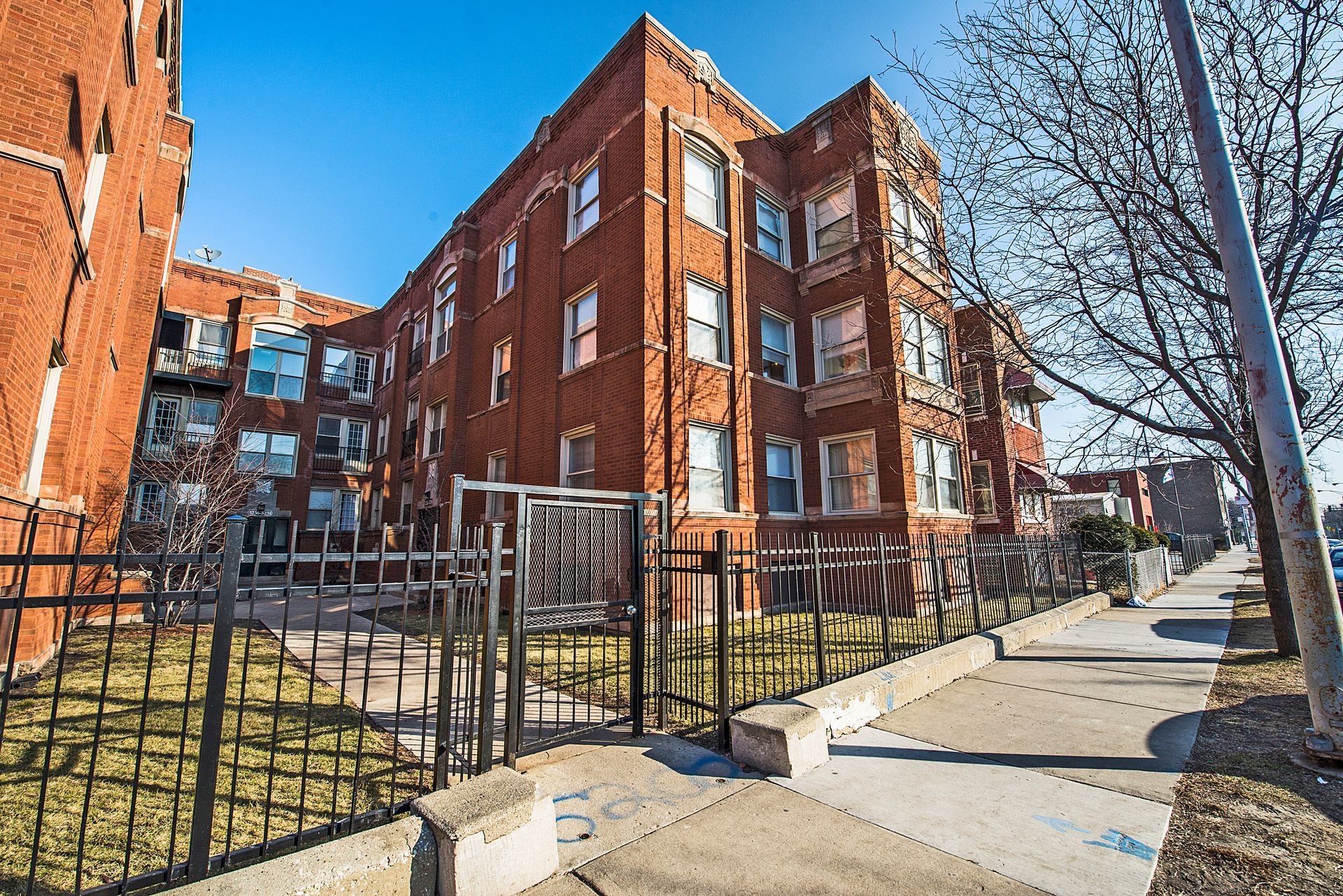 Red brick apartment building with black fence and sidewalk.