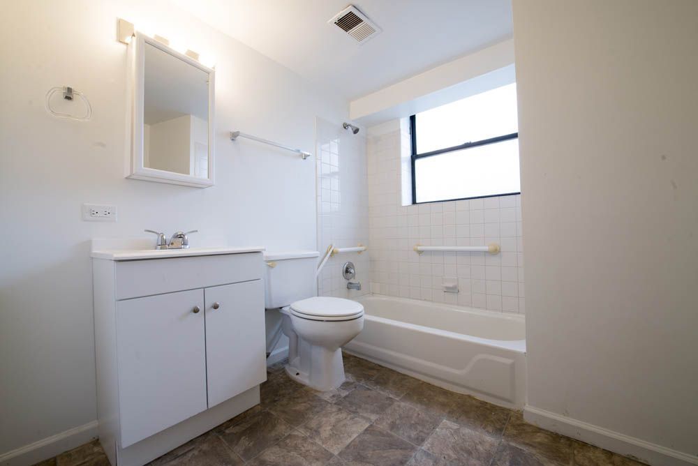 Bathroom with white fixtures, vanity, toilet, and bathtub. Gray floor tiles and a window.