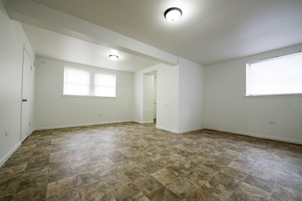 Empty, well-lit room with two windows, a doorway, and brown tile flooring.