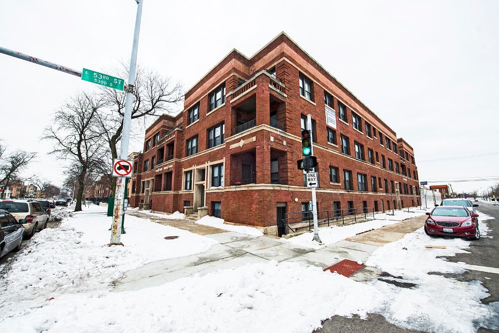 Brick apartment building at a snowy street corner with traffic light and sign. Cars parked along the street.