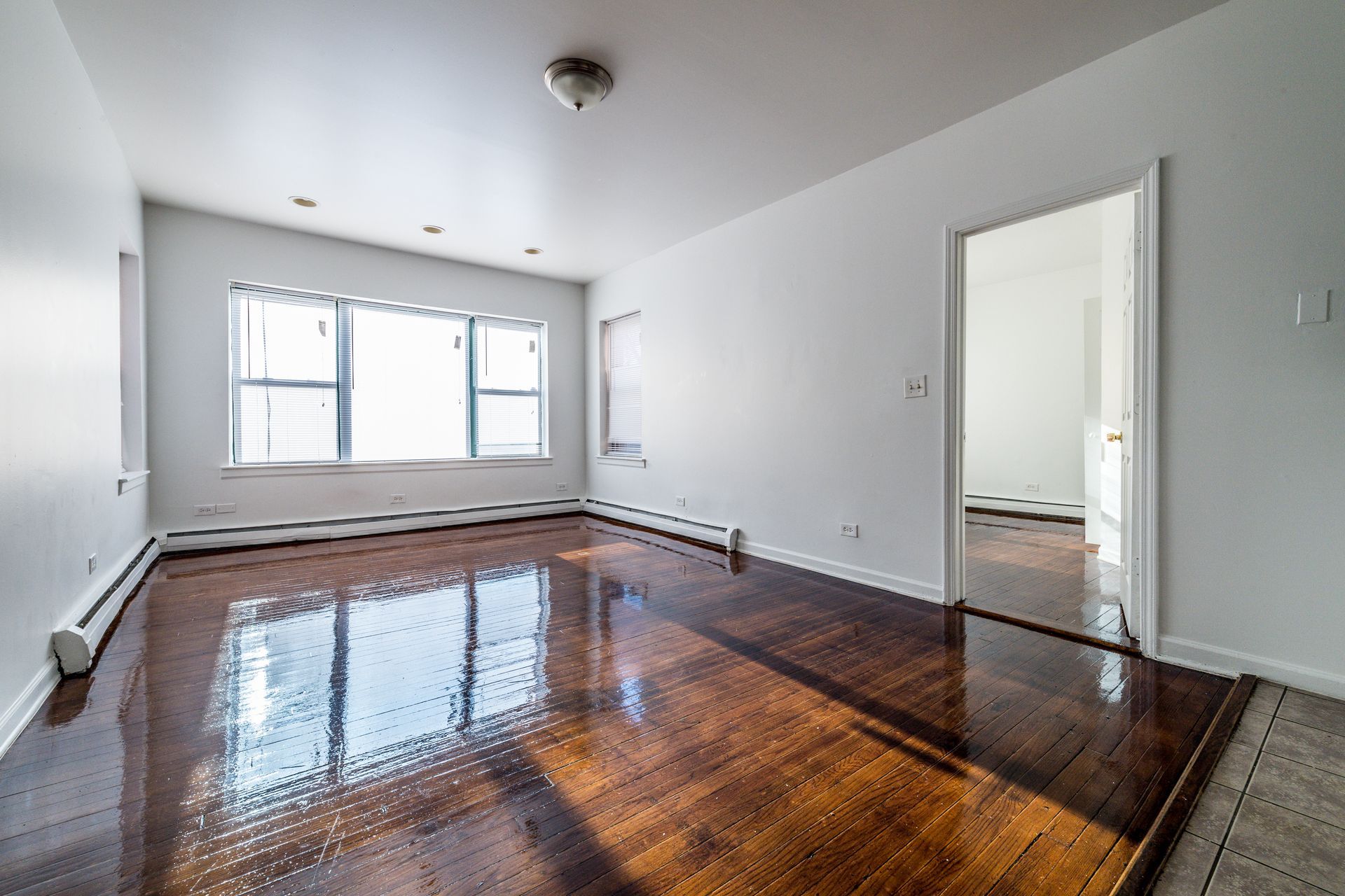 Empty room with shiny wood floors, large window, and open doorway. White walls, recessed lighting.