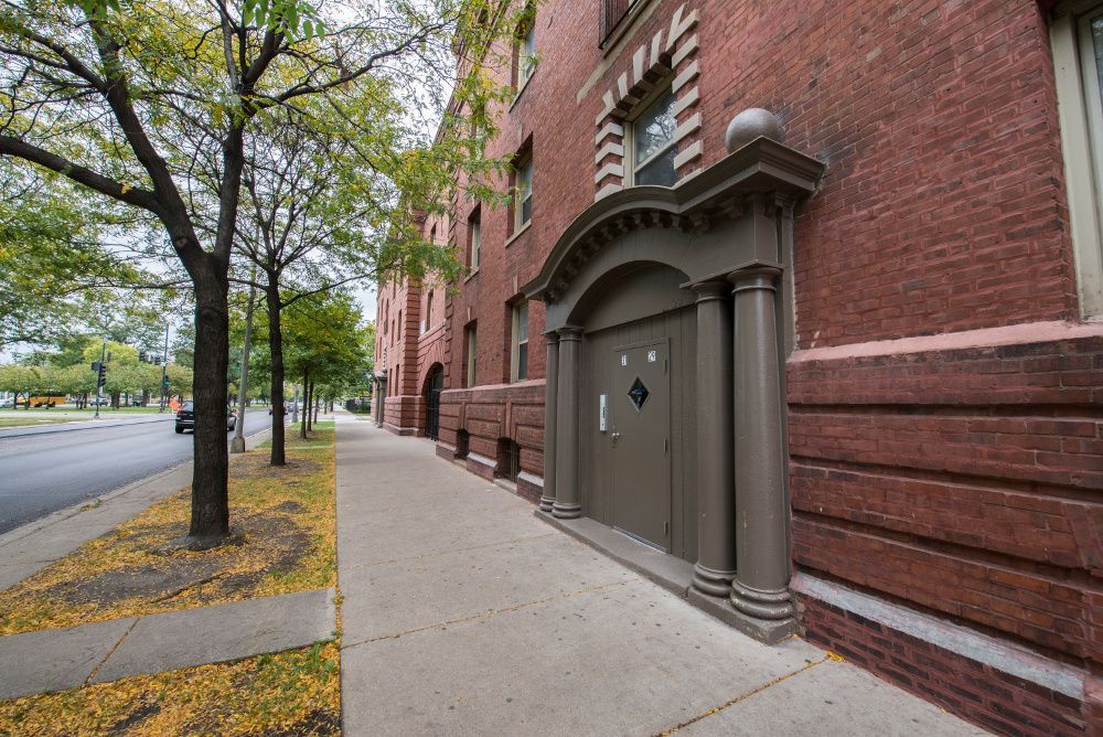Brick building with ornate door and sidewalk along a street, with a tree in the foreground.
