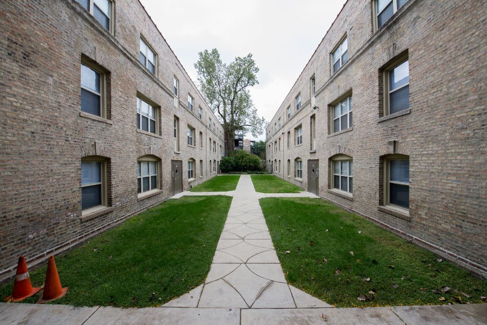 Two brick apartment buildings face each other, with grass and a walkway between them. A tree is visible at the end.