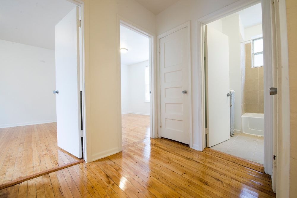 Hallway with hardwood floors, three doorways, two open. White walls, doors. Sunlight enters through windows.