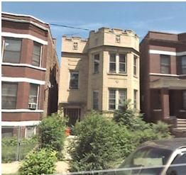Beige multi-story building with a turret, between two brick buildings, with shrubbery in front.