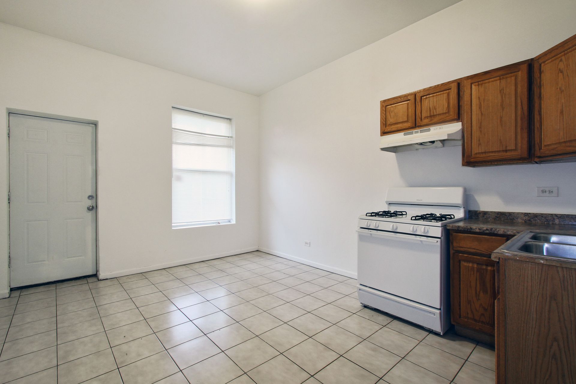Empty kitchen with white walls, cabinets, appliances, and tile floor; window and door on opposite sides.