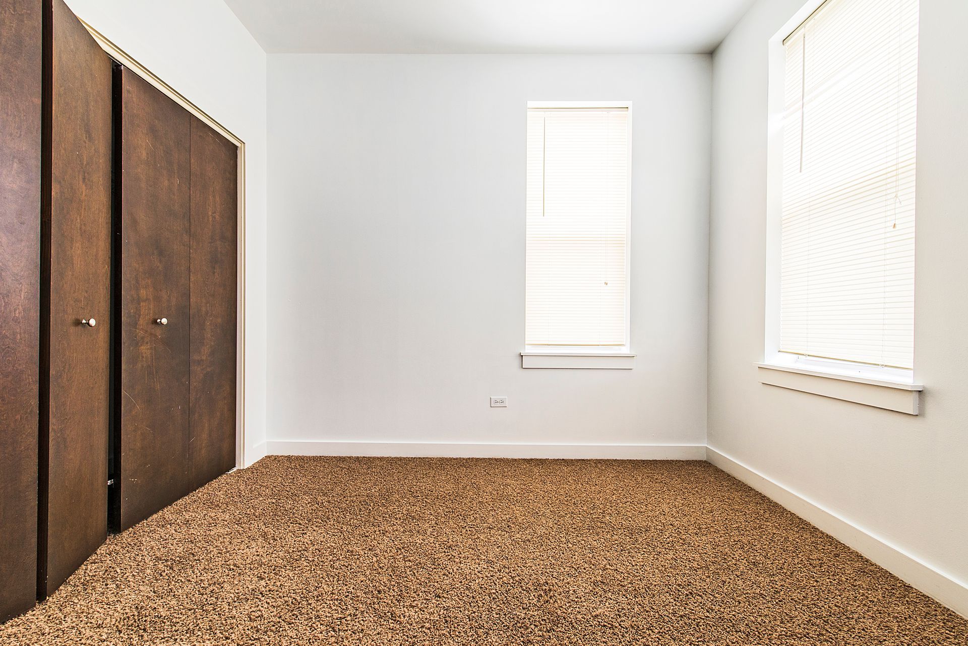 Empty room with brown carpet, white walls, closet, and two windows with blinds.
