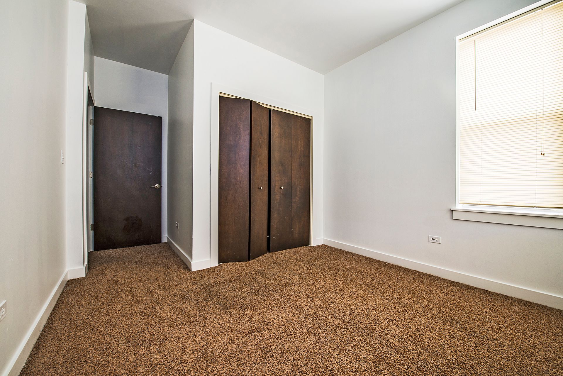 Empty bedroom with brown carpet, white walls, dark wood closet doors and a window with blinds.