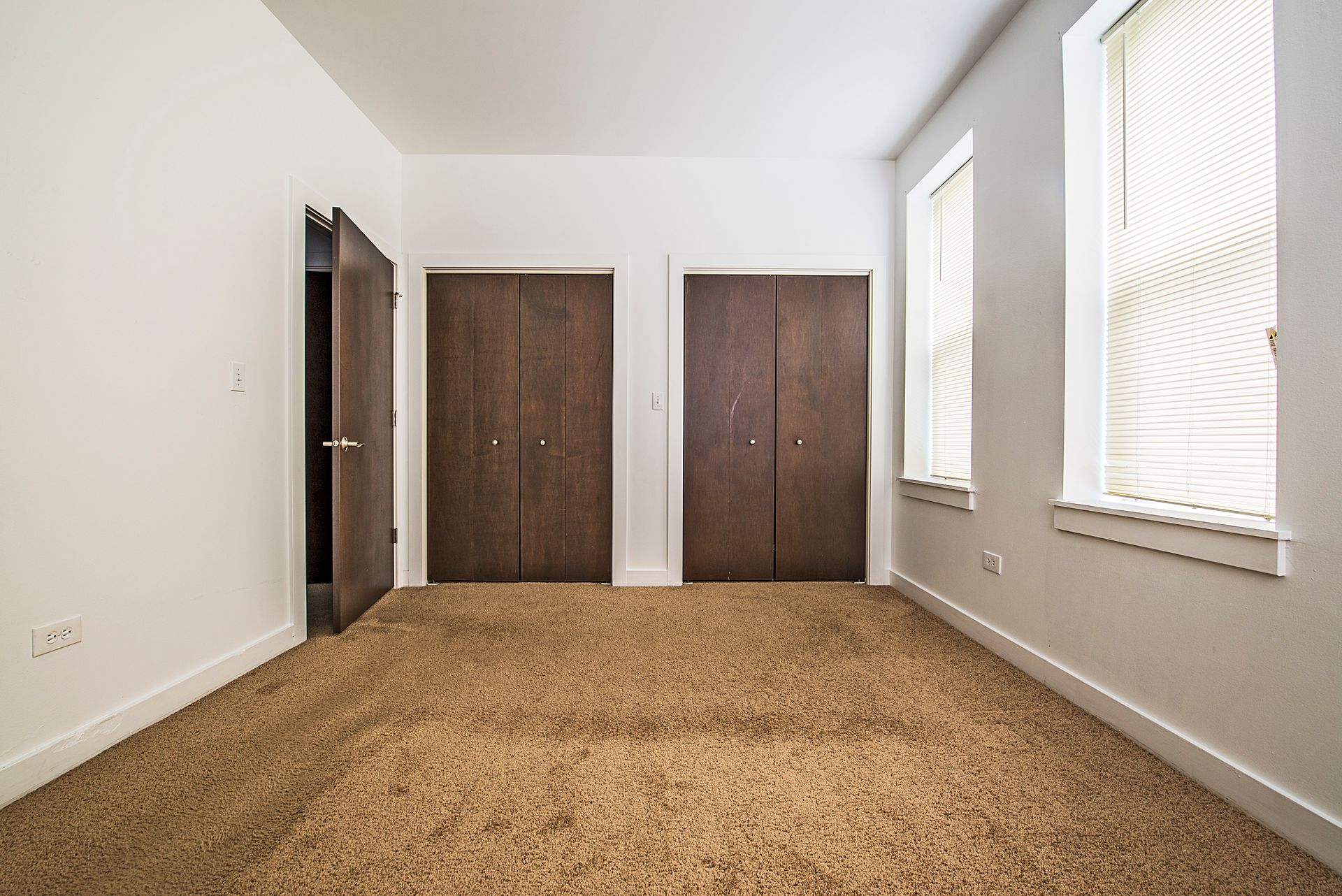 Empty room with brown carpet, two wood doors, two windows with blinds, and white walls.