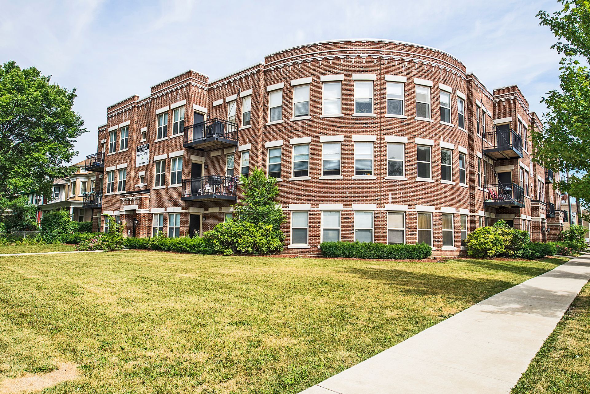 Brick apartment building with arched section and balconies, on a grassy lot, sidewalk along the right.
