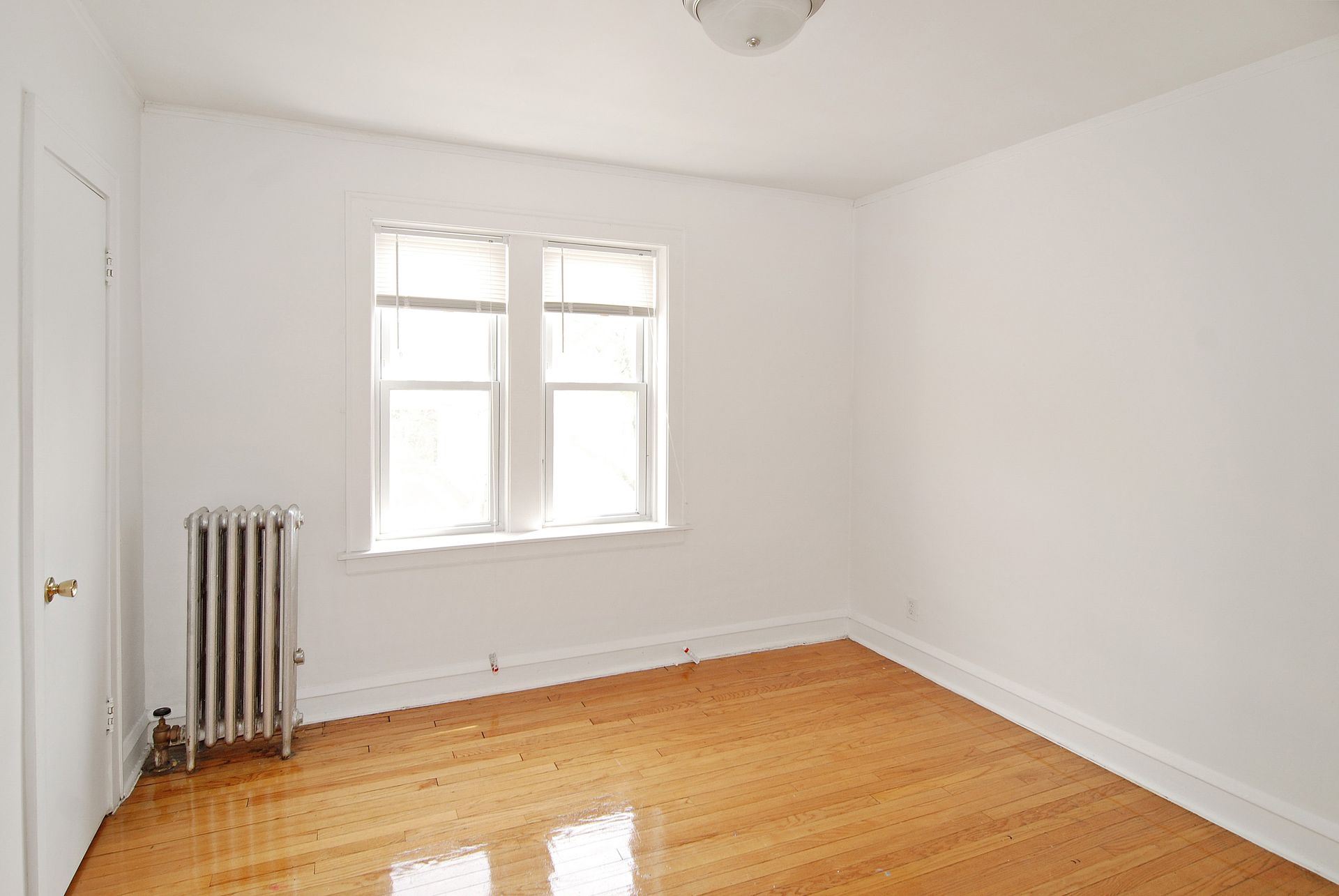 Empty room with wood floor, white walls, window, radiator, and a closed door.