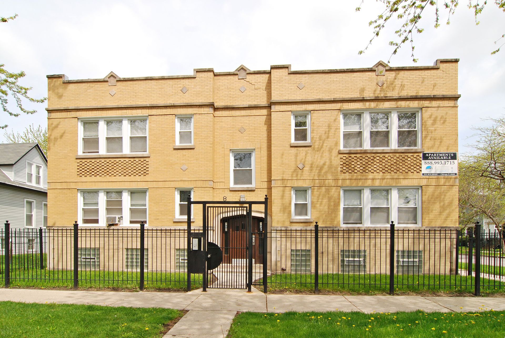 Two-story tan brick apartment building with black iron fence and gated entrance.