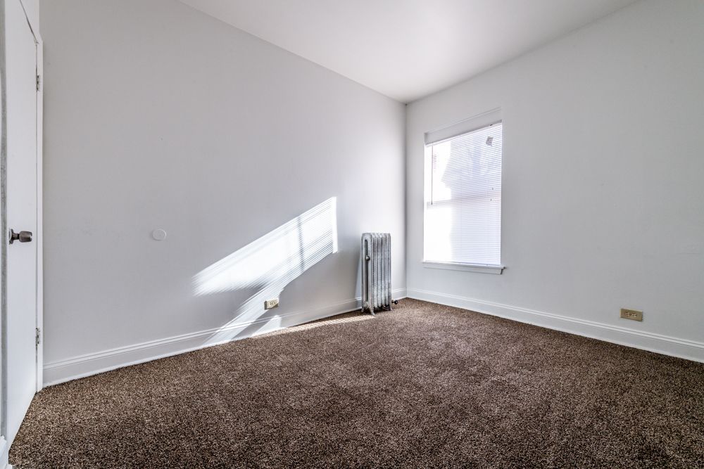 Empty room with brown carpet, white walls, window with blinds, and a radiator.